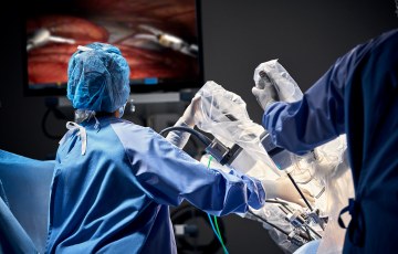 Leikkaussalissa sairaalan henkilökuntaa ja leikkausrobotti leikkaamassa potilasta taustallaan iso tv-ruutu, josta näkyy, kun instrumentit työskentelevät.