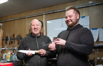 Kaksi miestä seisovat verstaalla ja pitävät kädessään käsintehtyjä keittiöveitsiä