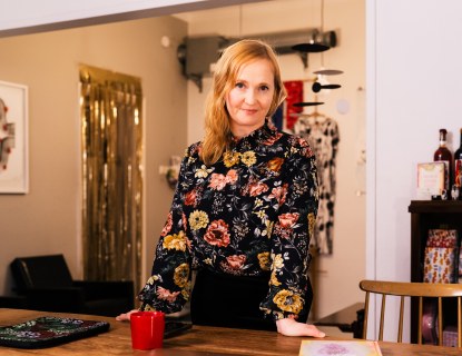 Nainen seisoo pöydän takana ja nojaa siihen kodinomaisesti sisustetulla toimistolla