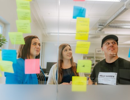 Kolme ihmistä toimistohuoneessa ison ikkunan edessä. Ikkunassa on kiinni postit-lappuja. 