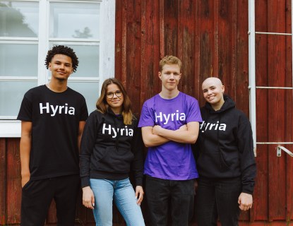 Neljä nuorta seisoo rivissä, punaisen talon edustalla. Heillä on päällään paidat jossa on Hyrian logo.