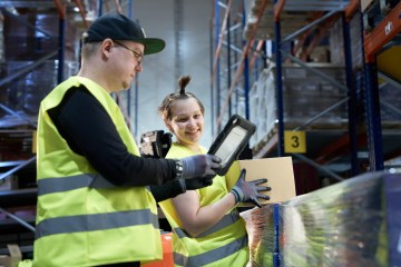 Kaksi varastotyöntekijää keltaisissa liiveissä katselevat tabletin näköistä laitetta, toisella pahvilaatikko kädessään.