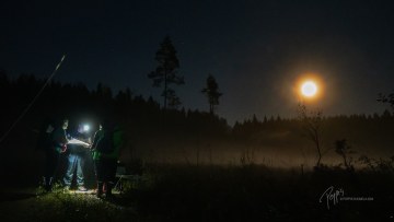 Retkeilijöitä pimeässä metsässä otsalamppujen ja kuun valossa tutkimassa karttaa