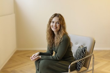 Valokuva jutussa esiintyvästä Aino Kiurusta. Aino istuu tuolilla tyhjässä huoneessa, ja hymyilee kameraa kohti. Hänellä on päällään tummanvihreä mekko. 