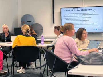 Henkilöt istuvat pöytien ääressä ja keskustelevat